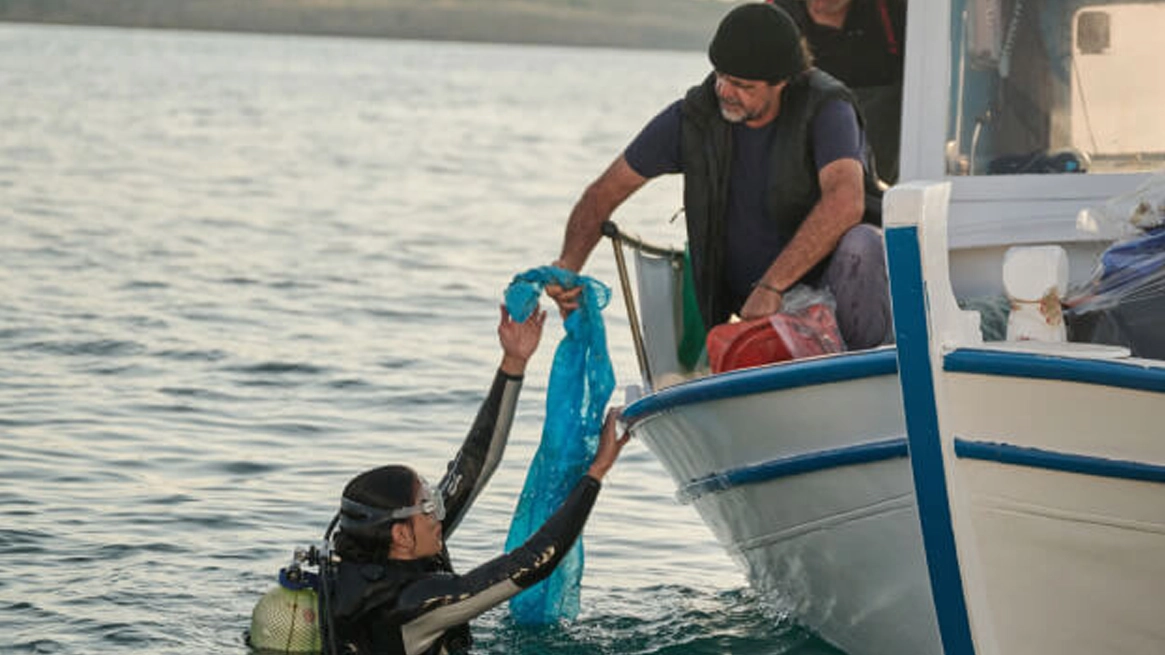The image depicts a maritime rescue situation, likely involving a boat crew assisting someone in distress out of the water. Two individuals are interacting with each other: one is reaching towards the water to retrieve an object that appears to be floating in or near the water, while the other person nearby inside the vessel might be preparing to assist the individual being rescued.