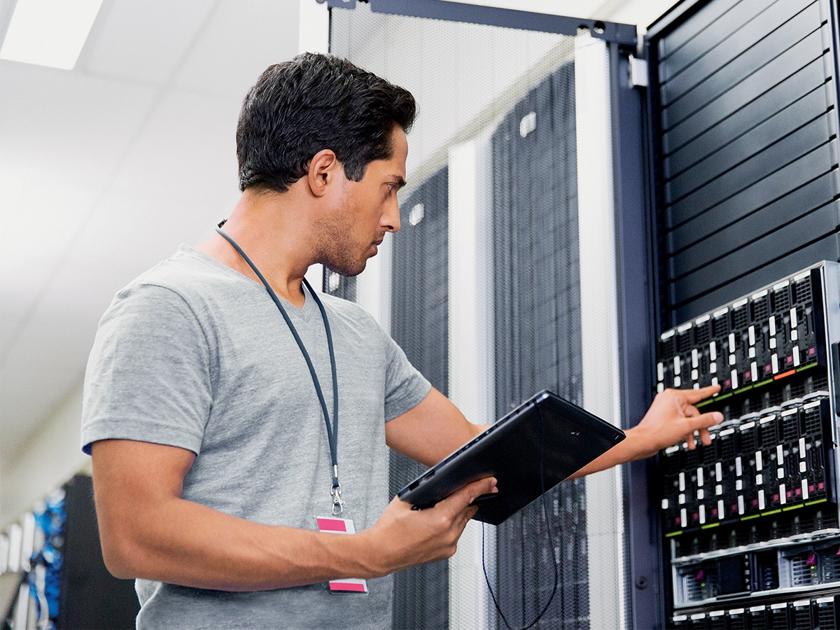 In this image, a person is standing next to what appears to be a server rack in an IT or data center environment. The individual is holding a tablet and seems to be operating it.
