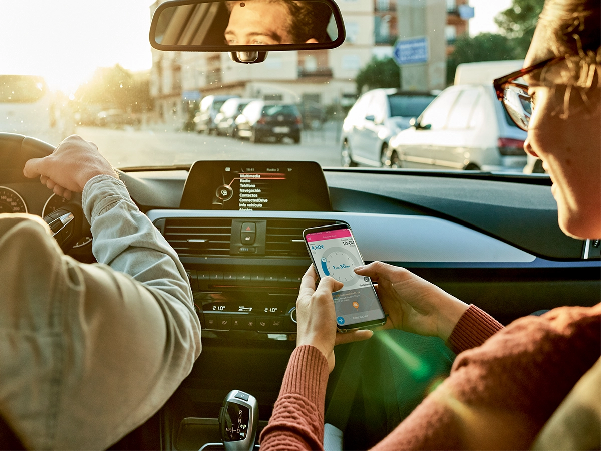 The image shows two people in a car, with the man in the driver's seat looking to his left. The woman, sitting in the passenger seat, has her attention on her phone, which she is holding with both hands.