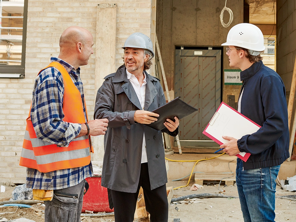There are three individuals at what looks like a construction site. There are three men in the foreground, and they seem to be engaged in conversation or a discussion. Two men are holding clip boards and wearing helmets.