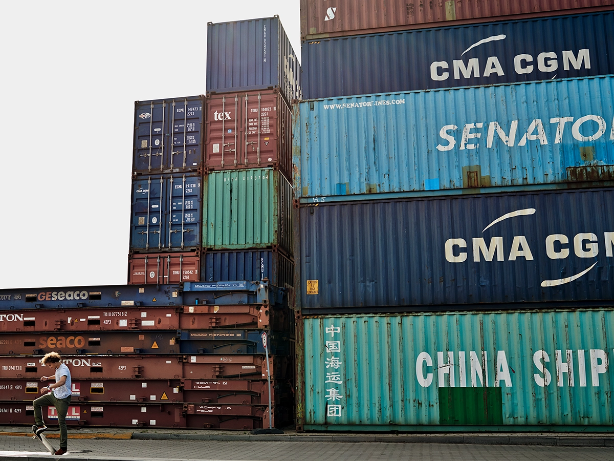The image shows a scene of an urban setting with shipping containers stacked in the background. In the foreground, there's a person doing tricks on a skateboard.