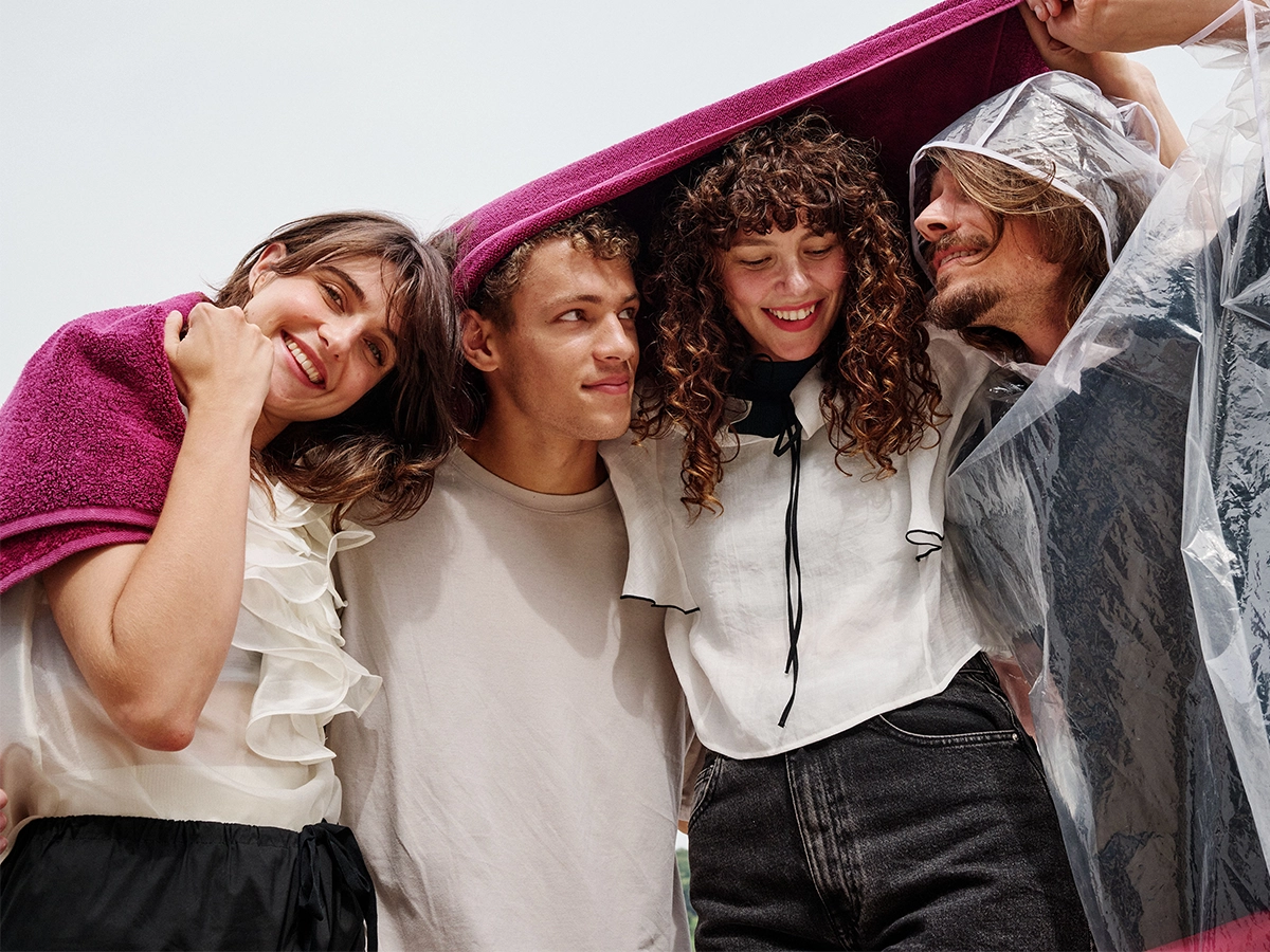 In the image, there are four individuals in an outdoor setting. The group appears to be enjoying a moment together under a towel, which they hold up together.