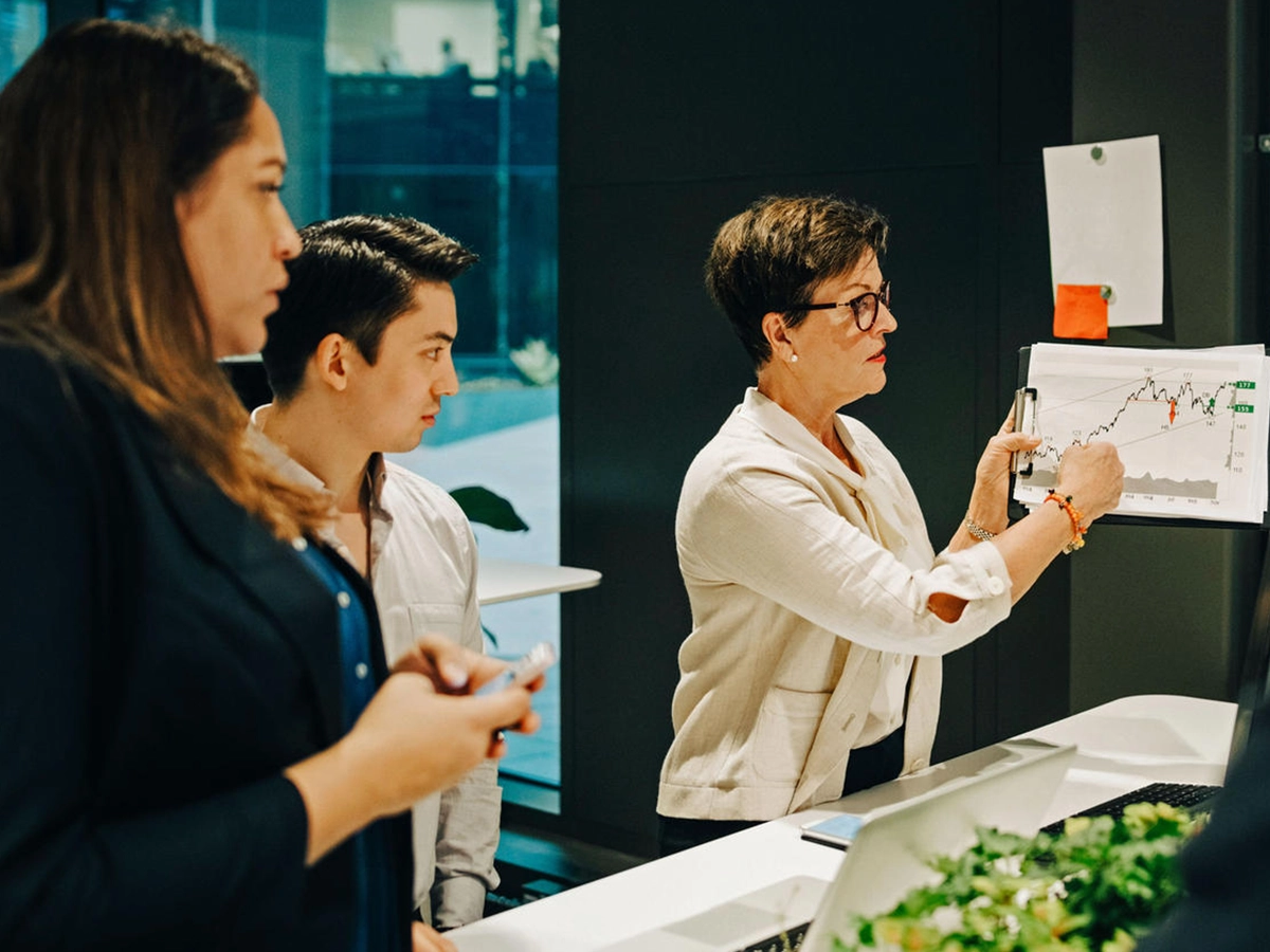 This image shows a person presenting or explaining a graph on a clip board, to at least two other people. It seems like an office setting.