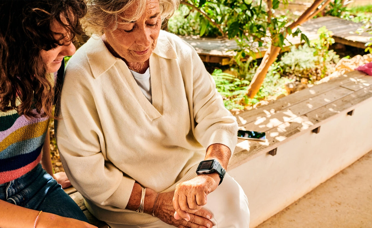One elder woman and another woman are sitting outside. They are both looking at the smart watch of the elder woman.