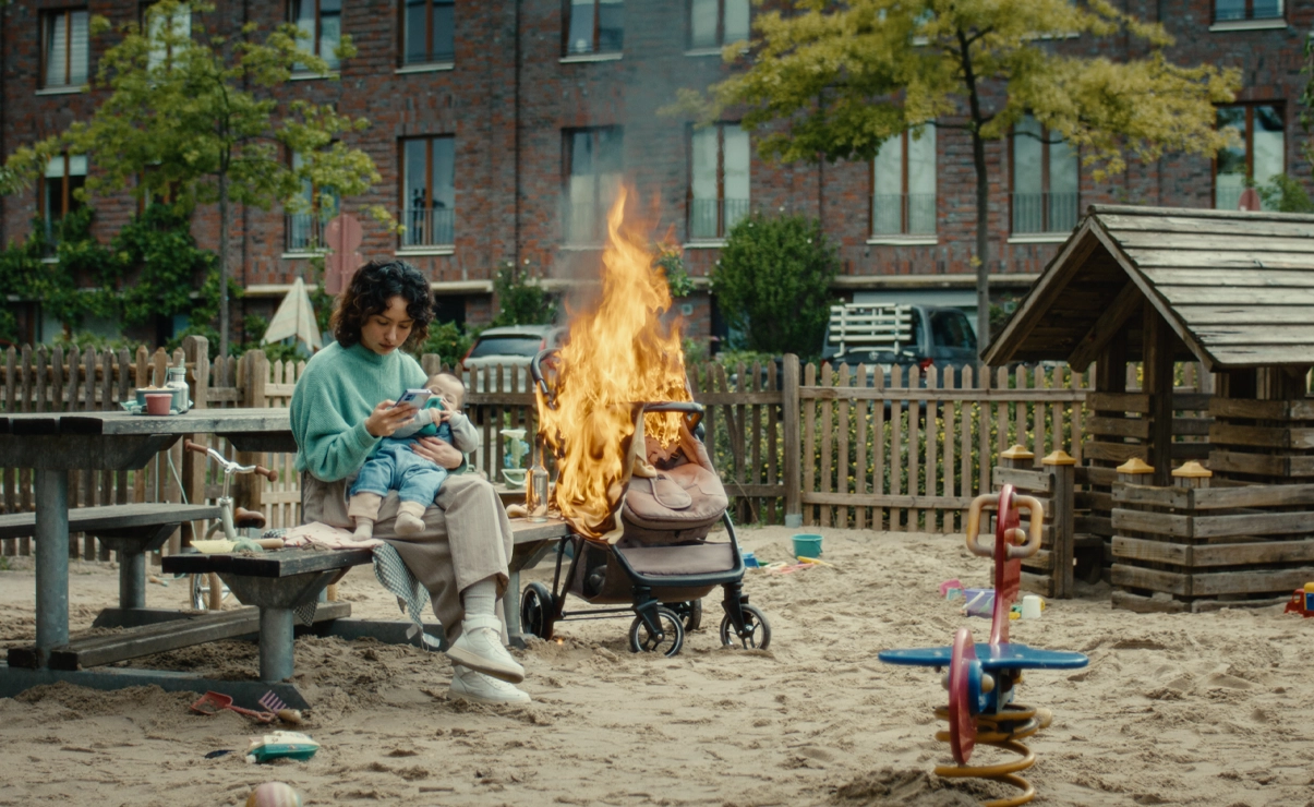 This picture is a scene from a film. It shows a person sitting on a playground, holding a baby and looking at here phone. Behind here the stroller is burning.