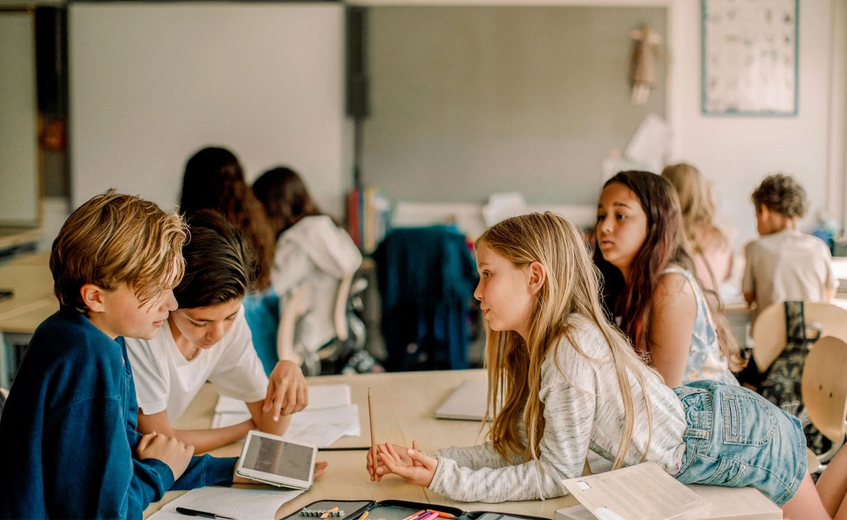 It depicts a group of students in a classroom setting, focusing on their papers or screens. Some are seated at desks while others are standing around, collaborating or engaged with their studies.
