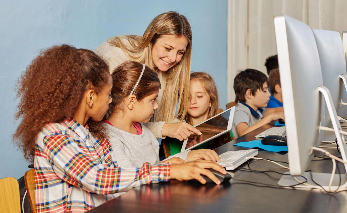 The image shows a classroom setting where a woman is teaching children how to use computers.