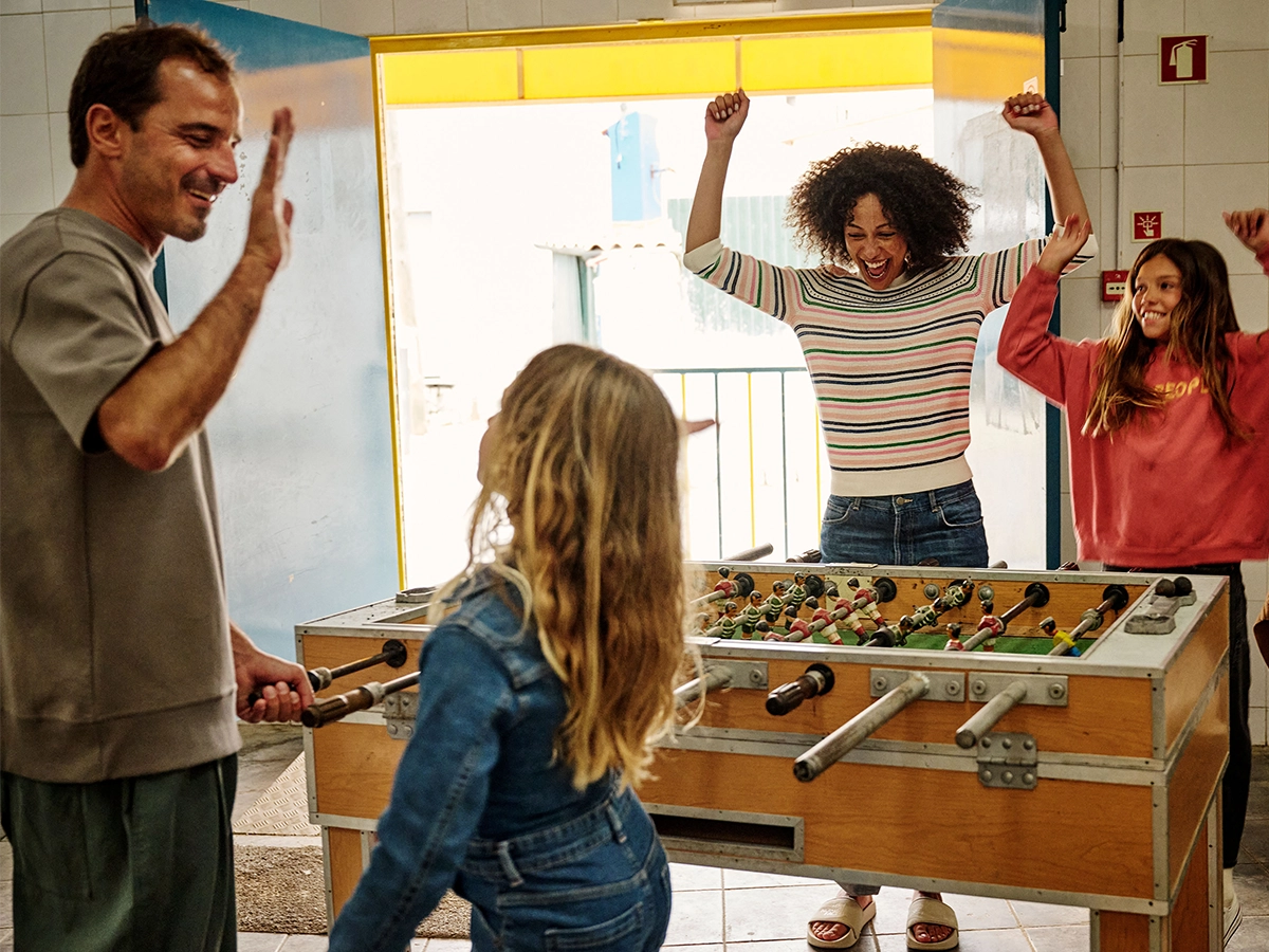 The image shows a casual indoor scene where a group of people, including both adults and a child, are gathered around a table containing two foosball tables. An adult male is showing off his skill in playing foosball, making a dynamic move with his hand raised above him. He appears to be the focus of attention as he interacts with the games. 