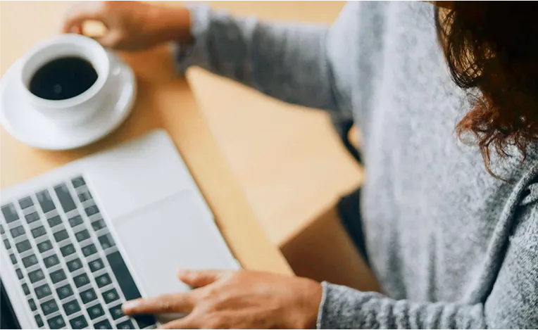 Woman in a grey pullover drinks coffee while working on a laptop (Photo)