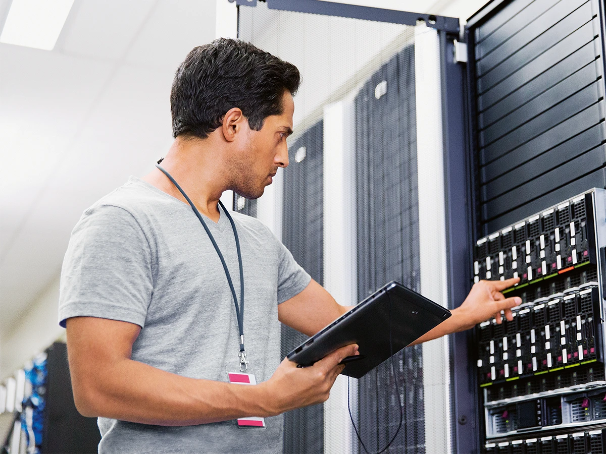 A man is standing in a server room, holding a laptop. (Header Photo)