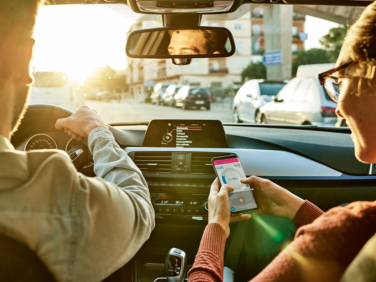 Two persons in a car. One person is steering while the other person is looking at a mobile phone. (Header Photo)