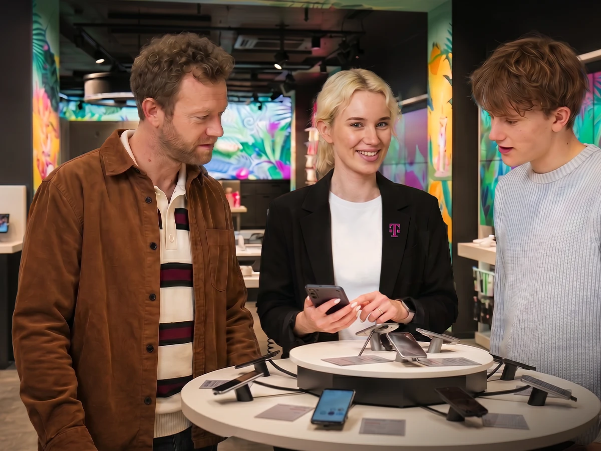 A woman with Telekom jacket shows two men a mobile phone in a Telekom showroom (Header Photo)