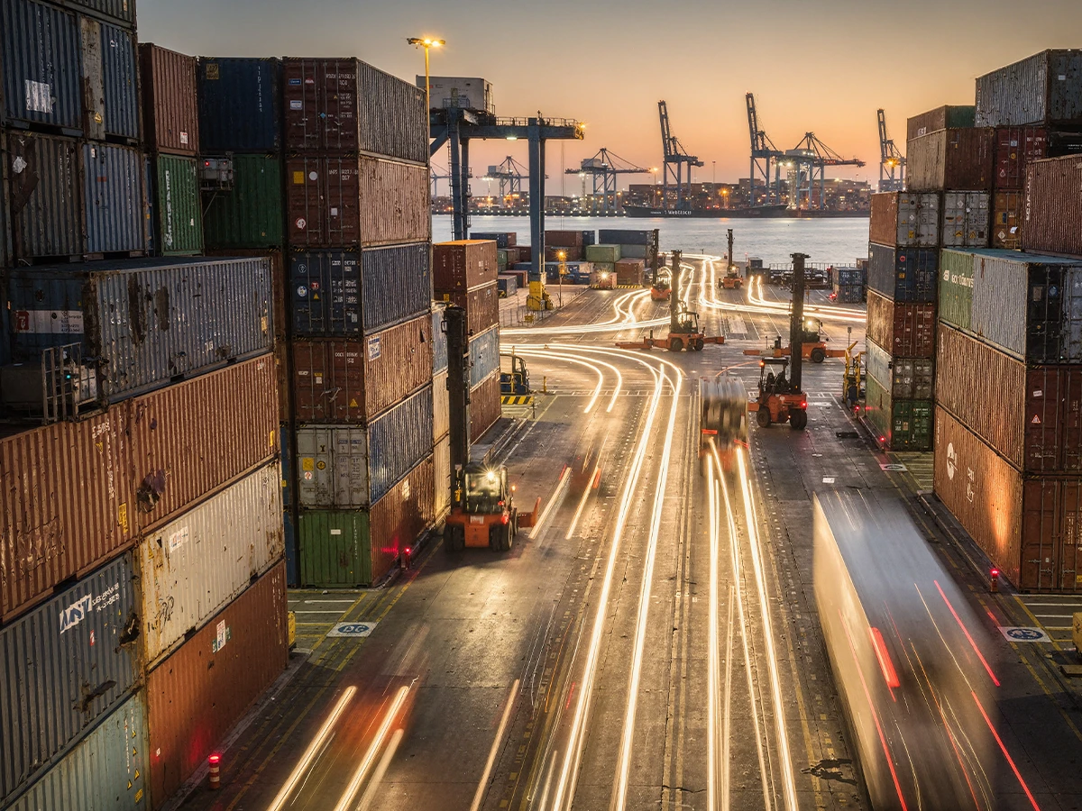 A dock at dusk or dawn. To the left and right are stacks of containers. Cranes are in the background and a road is running up the middle with streaks of lights from vehicles. (Header Photo)