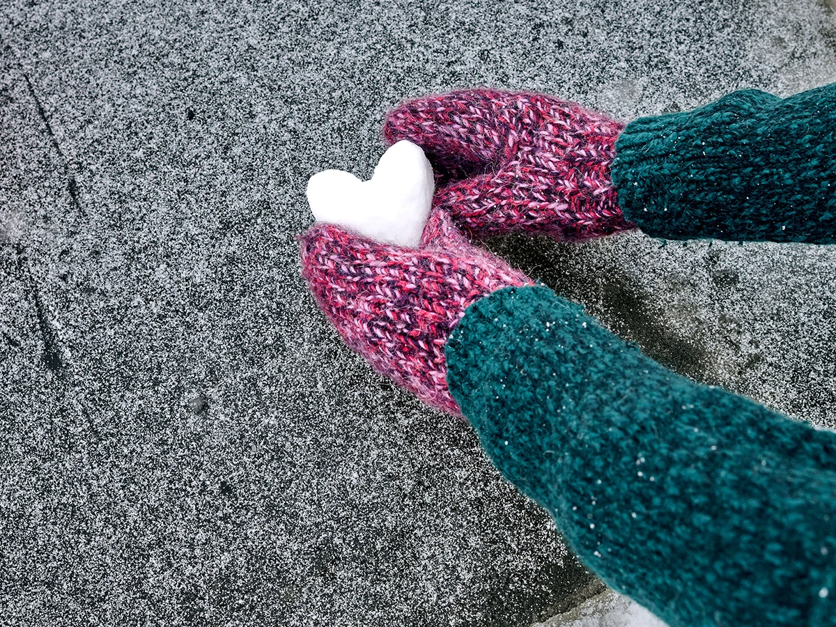 Close-up of someone forming a heart out of snow and wearing mittens. You can only see their arms and hands. (Header Photo)