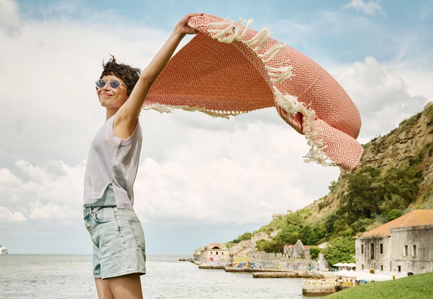 A woman wearing sunglasses is standing on a beach, holding an orange-patterned beach towel up to the wind. (photo)