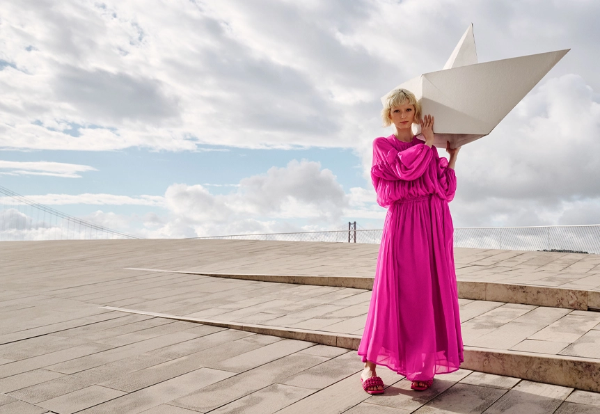A woman in a magenta colored dress holds an oversized paper boat on her shoulder. (photo)