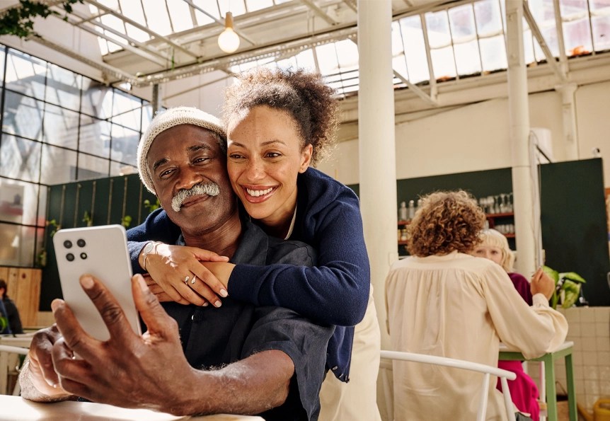 A man sits in a cafe and gets a hug by a woman. Both are looking at a mobile phone that he holds in his hand. (photo)
